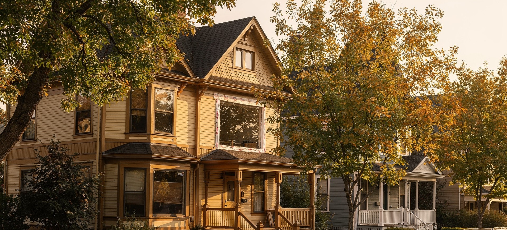 A tree-lined street of craftsman and victorian houses in Boise's North End in warm morning light, one home mid-renovation with a window opening carefully masked for replacement.