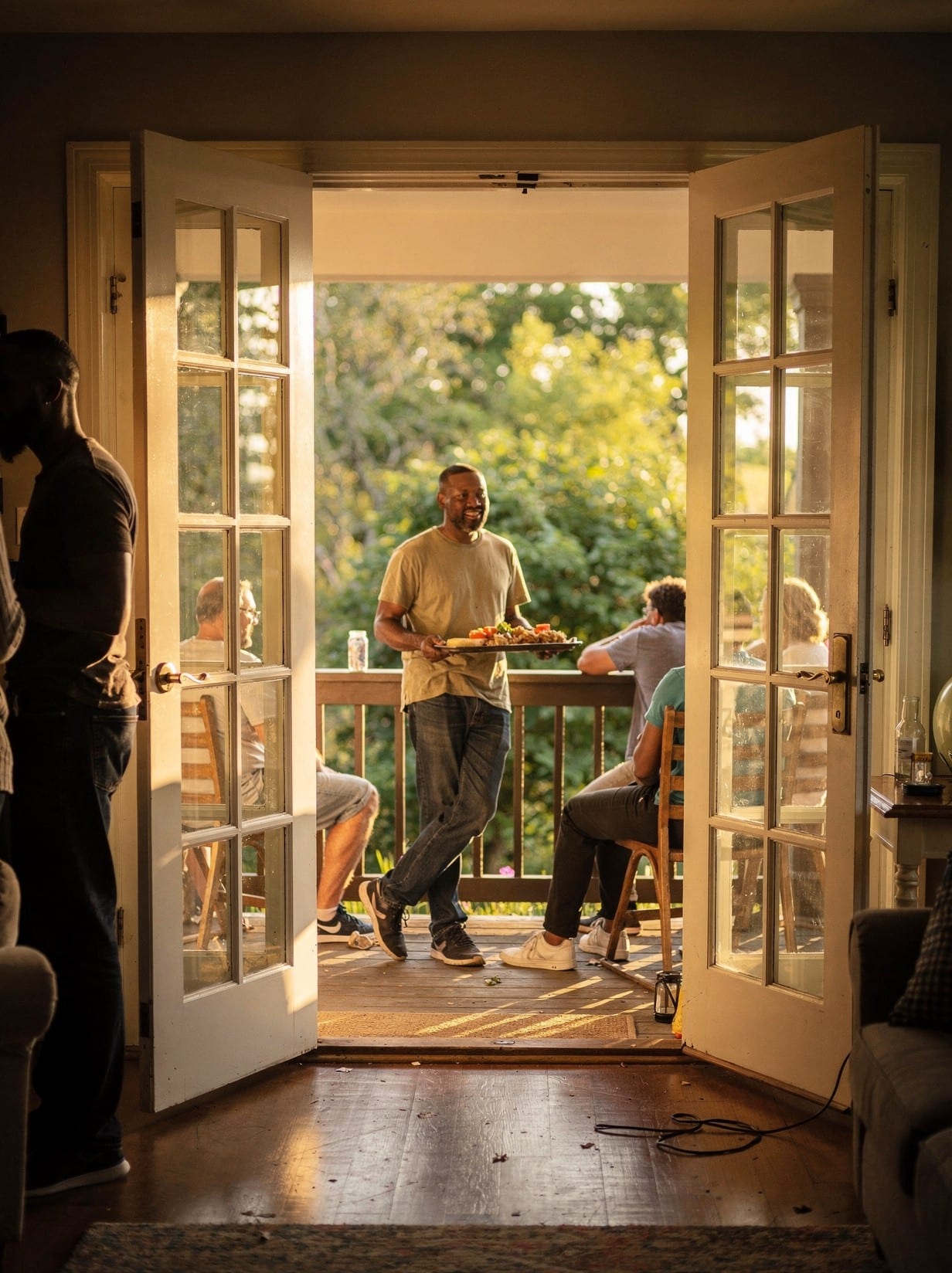 French patio doors thrown fully open onto a porch on a golden NC evening, the entire doorway clear, a gathering spilling between inside and out as someone carries a tray through the wide-open pair.