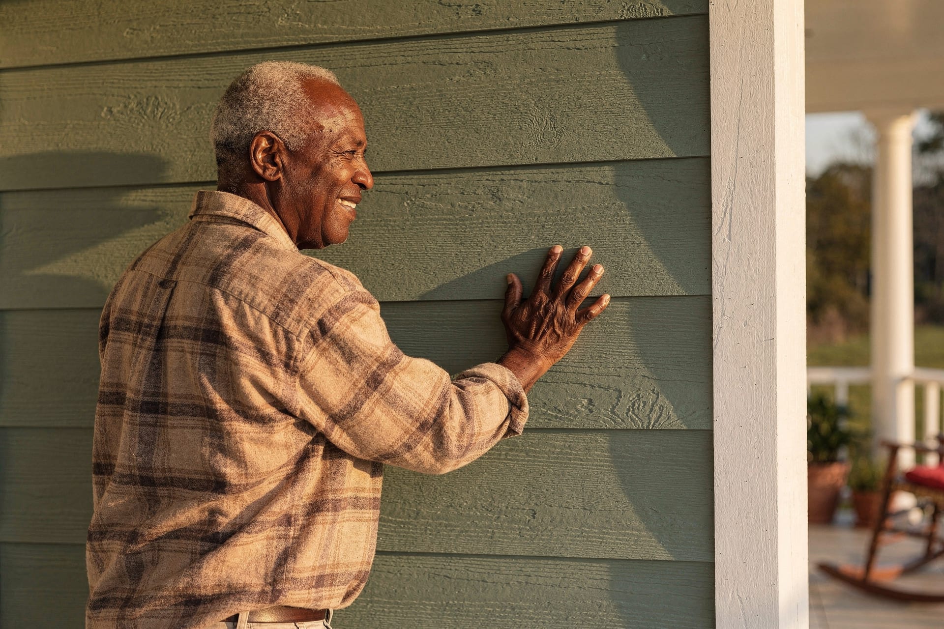 An older Black homeowner rests his hand on the deep wood-grain fiber cement lap siding of his North Carolina home, a quiet proud half-smile as warm afternoon light rakes across the wall he expects to outlast him.