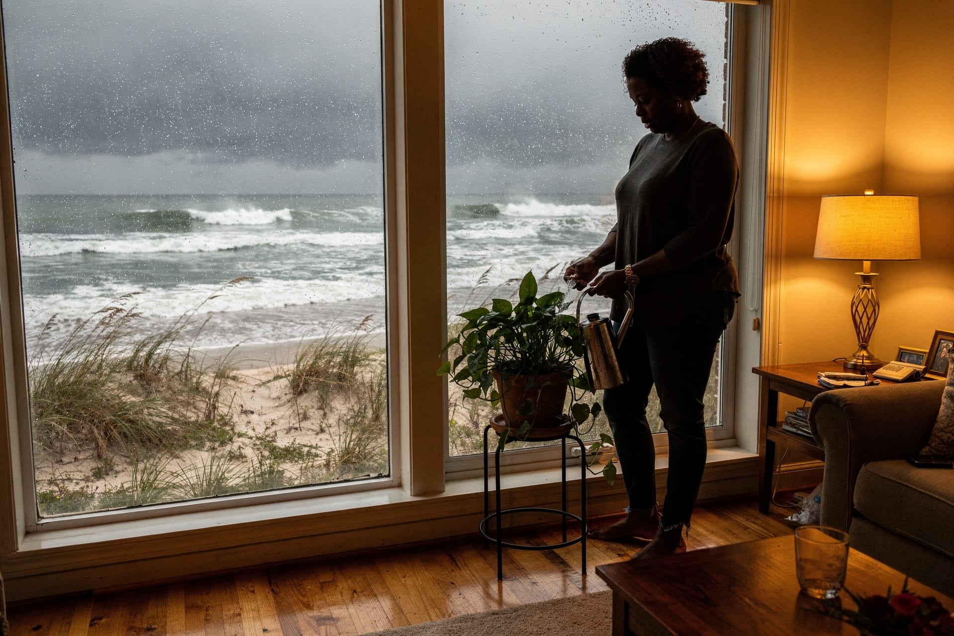 A homeowner calmly waters a houseplant beside a tall impact window in a warm coastal North Carolina living room while the first grey squall band of a storm churns the ocean and bends sea oats outside the glass.