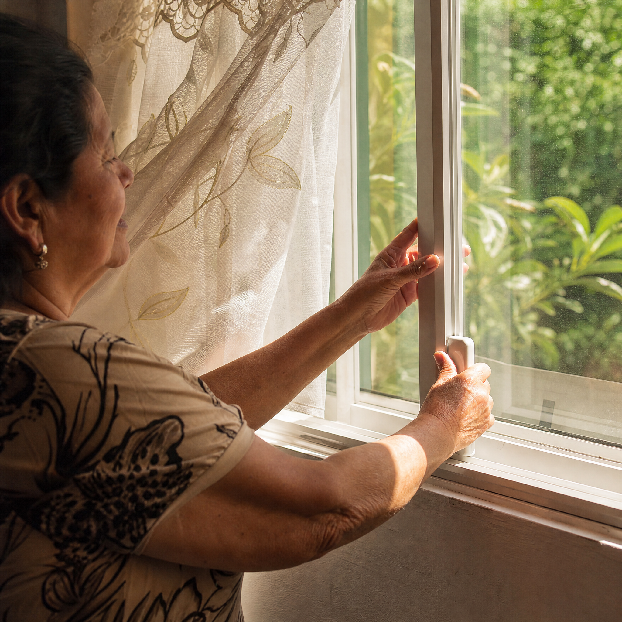 An older woman opening a new window with one hand in warm morning light, the sash gliding easily and a lace curtain lifting in the fresh air, green garden beyond the glass.