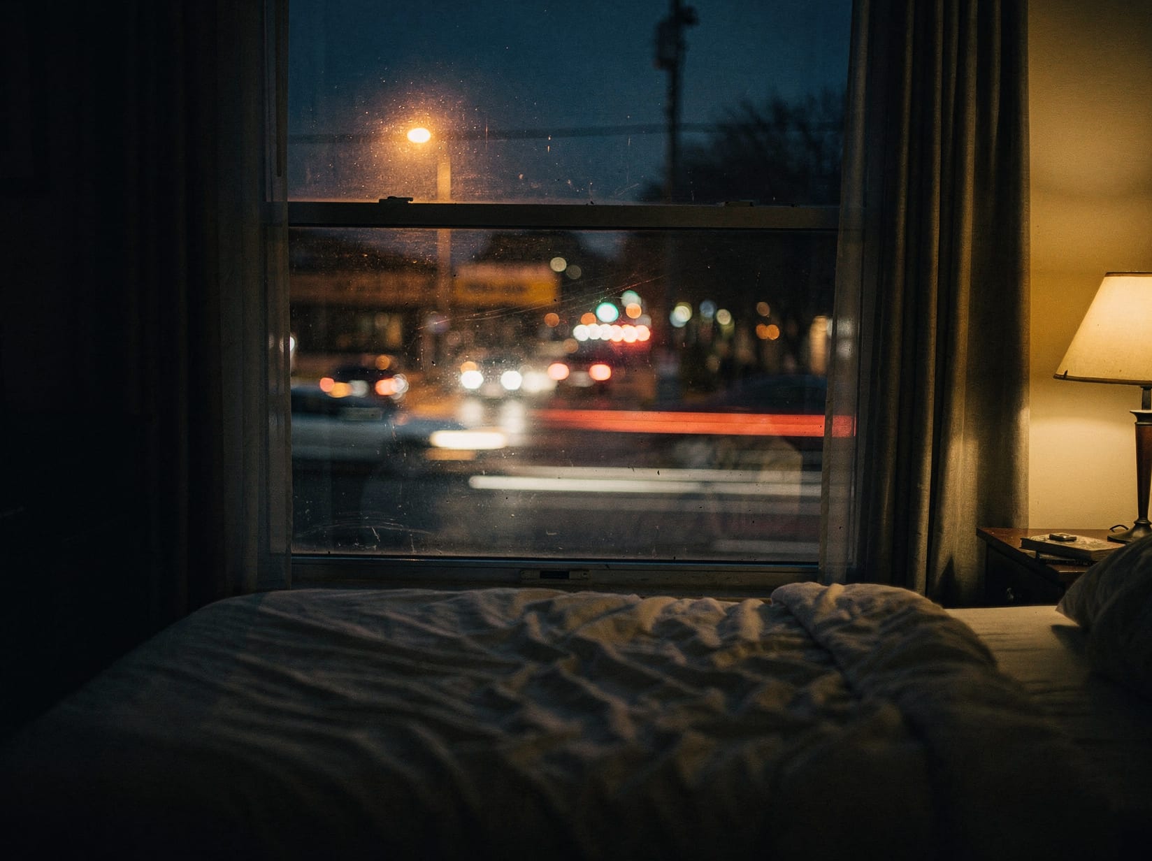A bedroom window at dusk facing a busy street, headlight streaks blurred through the glass and a streetlight glowing beyond, a warm lamp lit inside the quiet room.