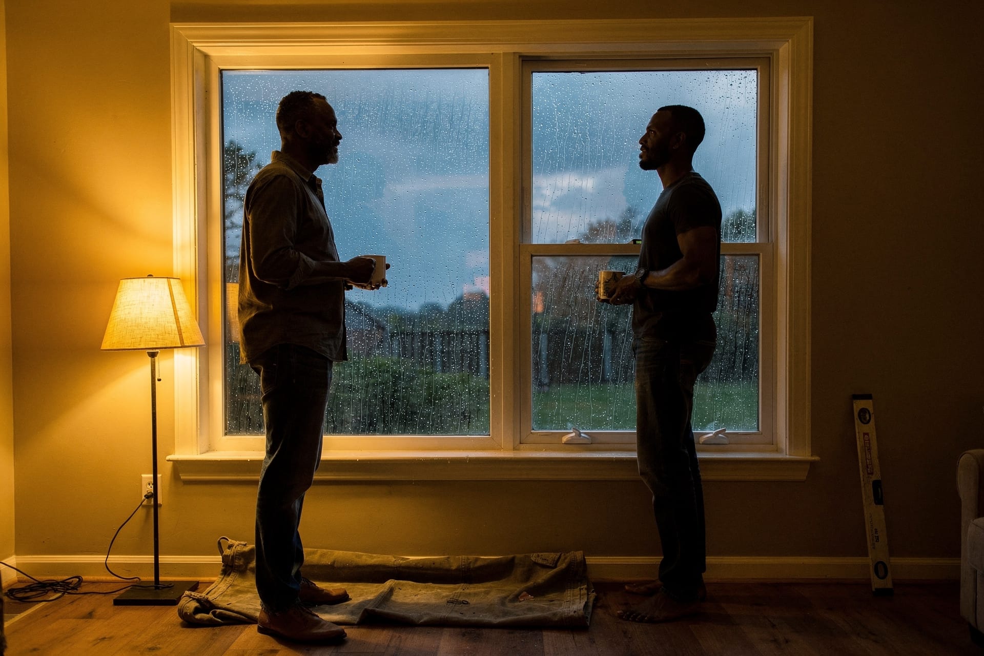 A homeowner stands relaxed beside a just-installed window in warm interior light while hard rain streaks the glass outside at dusk.