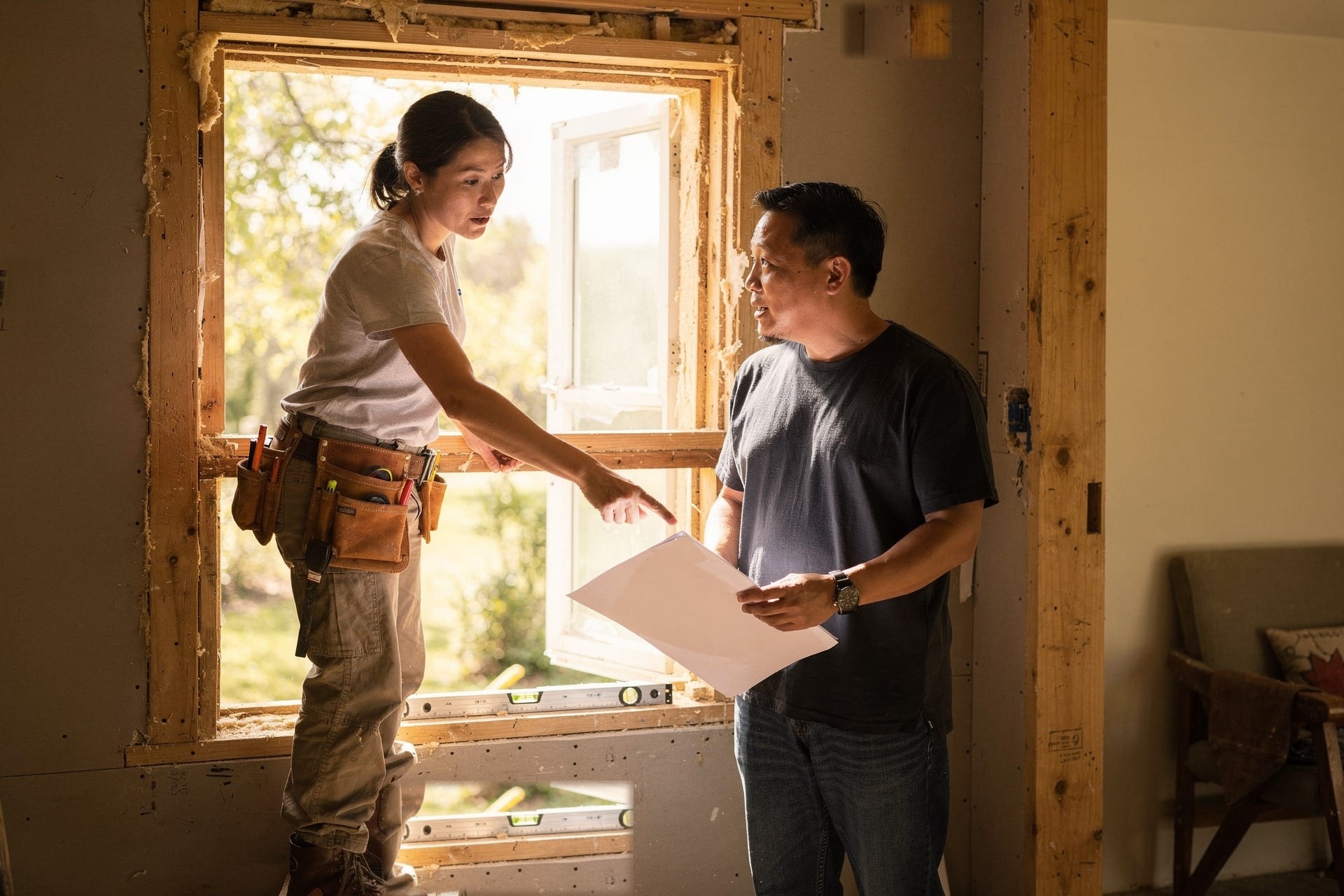 A homeowner holding a printed quote listens as an installer points at the exposed sill of an open window rough opening in daylight.