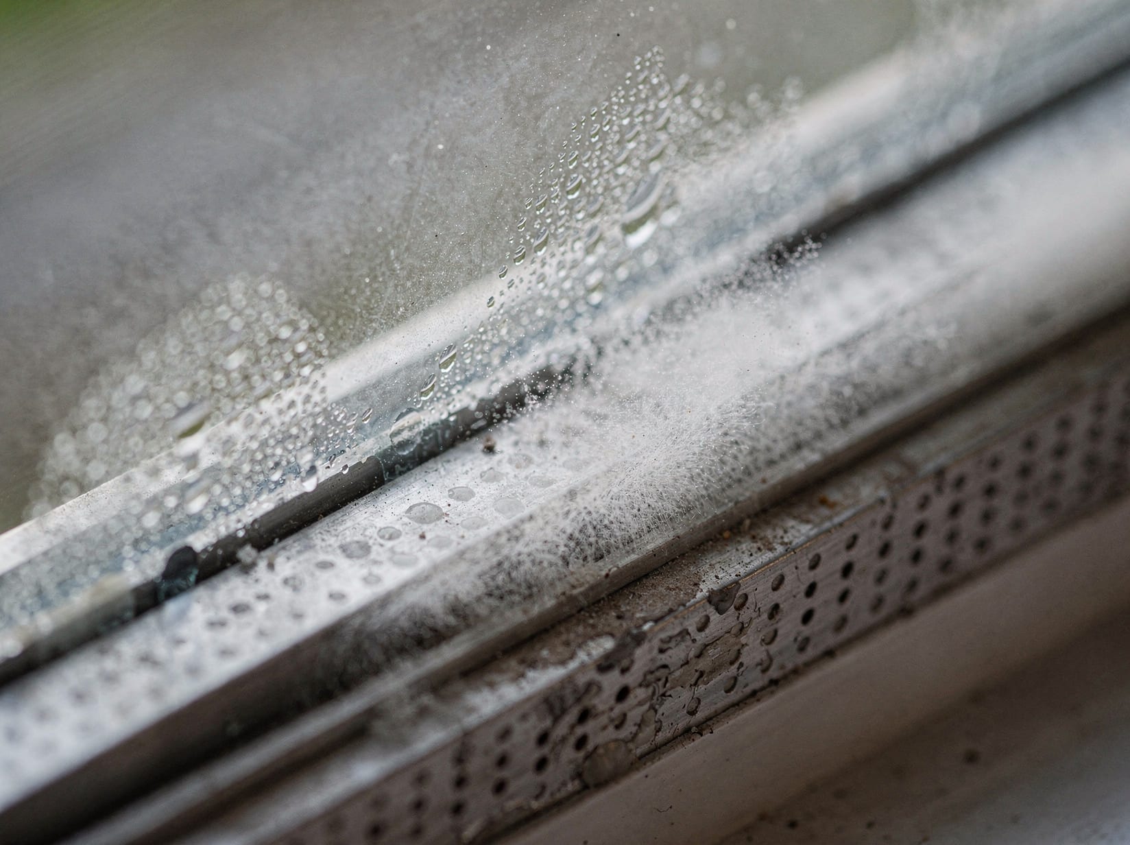 Extreme close-up of cloudy condensation droplets and mineral haze trapped between the panes at the glass edge near the spacer bar.
