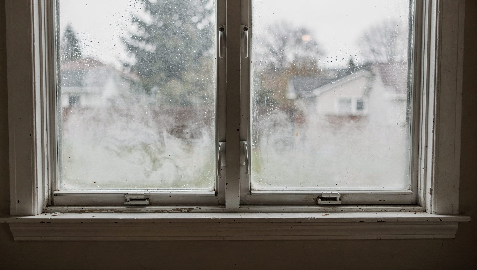 A residential double-pane window with cloudy condensation fog trapped between the panes and a hand resting a cloth flat against the glass mid-wipe, seen from inside under soft overcast light.