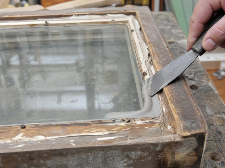 A wooden sash laid flat on a workbench being reglazed, a putty knife tooling a clean angled bead of fresh glazing putty into the rabbet along the edge of the glass, old putty stripped away and glazing points visible.
