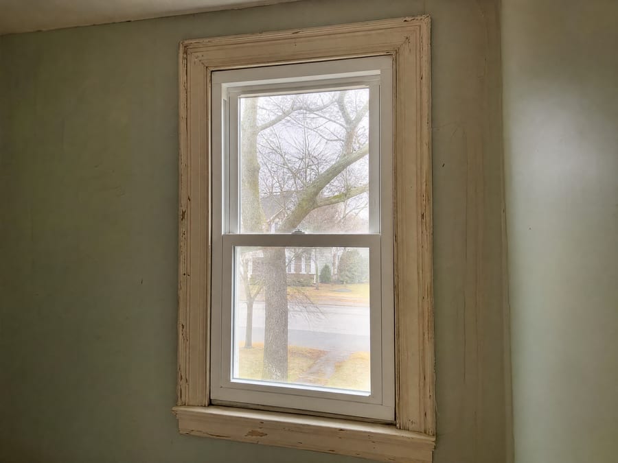 A crisp new white double-hung replacement unit fitted inside the opening of an older home, the chipped original painted trim and older plaster wall around it making the contrast between new insert and old house obvious.