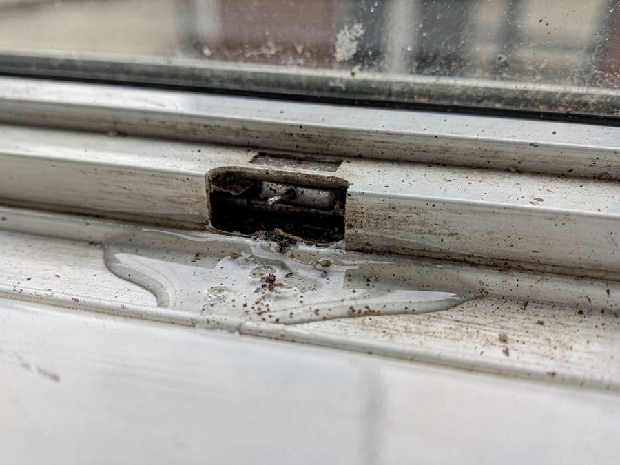 Macro of the bottom track of a white sliding window from inside, a shallow pool of rainwater standing in the channel and grit packed into the small drainage weep slot that should be letting it out.