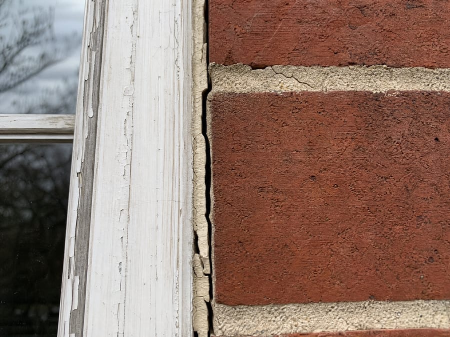 Exterior close-up of the joint where a weathered painted wood window frame meets red brick, the caulk bead cracked and pulled away leaving an open gap, with a hairline crack running through the mortar course beside it.