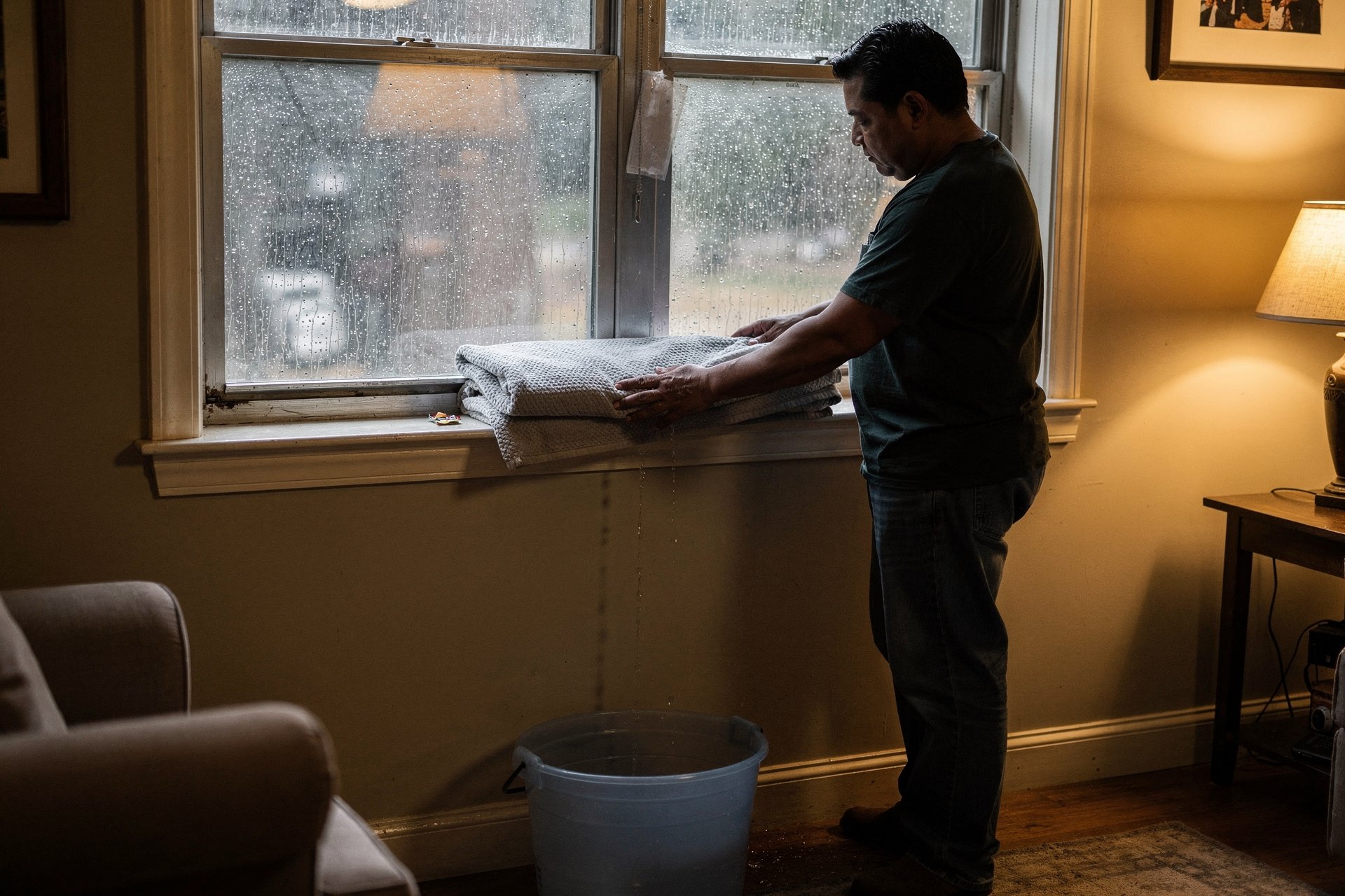 A Latino man in his late 30s calmly lays a folded towel along his living-room window sill and sets a bucket beneath a thin drip at the frame, while heavy rain streaks the glass outside — contained and capable, not panicked.