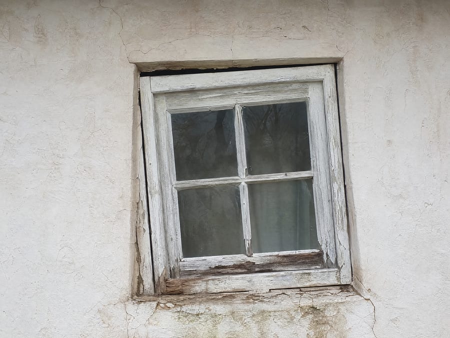 An old wooden window sitting visibly crooked in a settled opening, the head gap tapering to one side, long-term water staining and decay along the sill and cracks running through the wall around it.