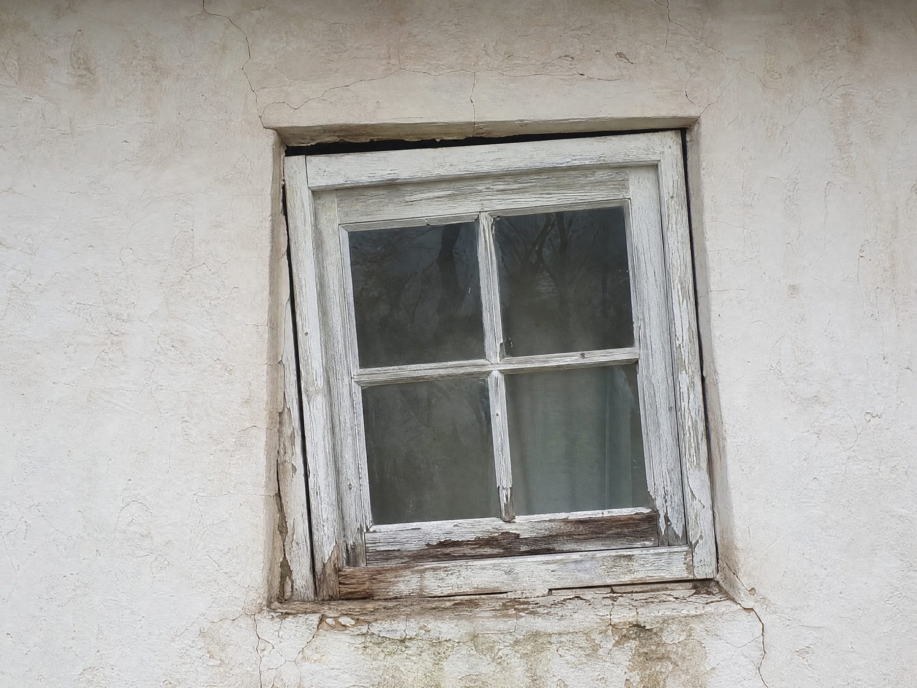 An old wooden window sitting visibly crooked in a settled opening, the head gap tapering to one side, long-term water staining and decay along the sill and cracks running through the wall around it.