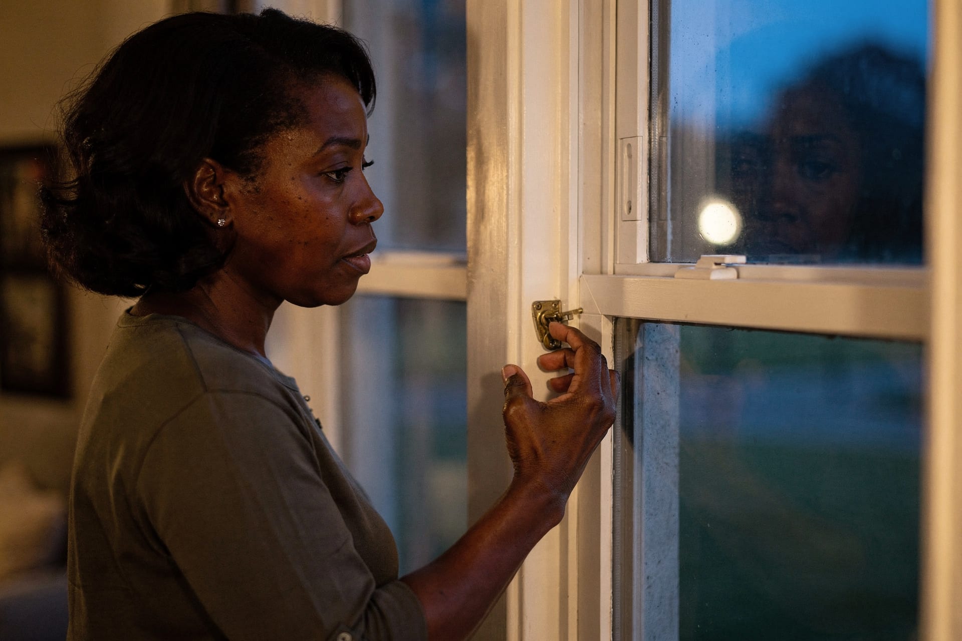 A woman stands beside a ground-floor double-hung window at dusk, one hand on the brass cam lock, testing a latch that won't quite catch — warm lamplight inside, blue evening beyond the glass.